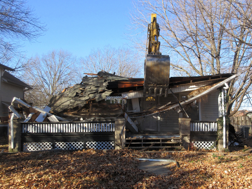 A hydraulic excavator crushing the roof of a house and crumpling the structure.
