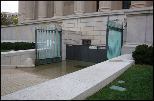 Automatic barrier installed across a large sub-ground level entry of a government building, with standing floodwaters visible.
