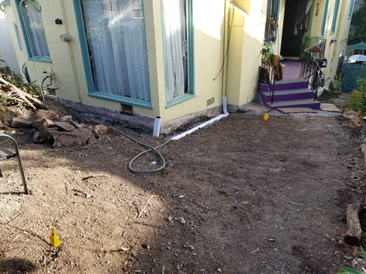 Trench dug around a house with a pipe installed waiting for gravel and topsoil. The pipe is connected to the downspout